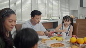 família feliz aproveita o café da manhã juntos. alegres pais asiáticos jovens e crianças pequenas bonitas desfrutam de café da manhã juntos na mesa de jantar em casa. video