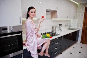 retrato de una bella joven vestida de rosa posando con un tomate en su cocina. foto