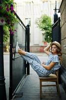 Portrait of a fabulous young woman in striped overall and hat sitting and posing on the chair outdoor. photo