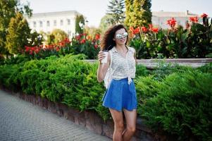 ropa de niña con estilo rizado en falda de jeans azules, blusa y gafas posadas en la calle de la ciudad con una taza de café. foto