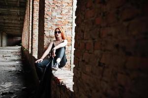 Red haired stylish girl in sunglasses wear in black, against abadoned place with brick walls. photo