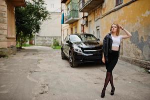 elegante chica rubia vestida con una chaqueta de cuero negro posando en las calles del coche de lujo de fondo de la ciudad. foto