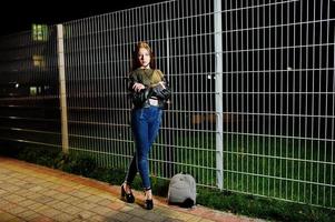 Night portrait of girl model wear on jeans and leather jacket against iron fence. photo