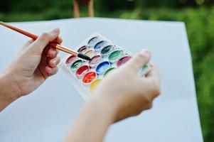 Close-up photo of female hands holding watercolor paints and a brush while painting in nature.