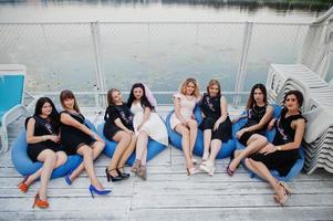 Group of 8 girls wear on black and 2 brides at hen party sitting on pillows at pier on beach side. photo