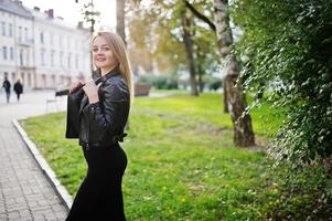 elegante chica rubia vestida con chaqueta de cuero negro posando en las calles de la ciudad. foto