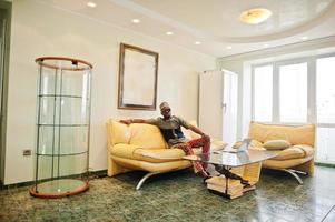 Rich african man sitting on sofa at his appartment and working on notebook. photo