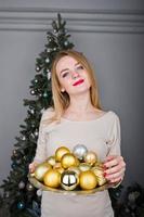 Cute blonde girl on beige dress against new year tree with with golden new year balls at studio. Happy holidays. photo