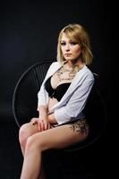 Studio portrait of sexy blonde girl with originally make up on neck and tattoo on thigh, wear on black underwear at dark background, sitting on chair. photo