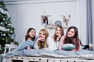 Four cute friends girls wear on warm sweaters and black pants on bed at new year decorated room on studio, play with pillows. photo