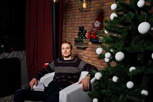 retrato de estudio de un hombre con un libro sentado en una silla contra un árbol de navidad con adornos. foto