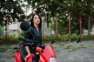 retrato de una mujer genial e impresionante con vestido y chaqueta de cuero negro sentada en una moto roja genial. foto