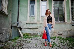 chica pelirroja con bolso rojo posó en la calle de la ciudad. foto