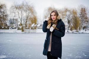 Beautiful brunette girl in winter warm clothing. Model on winter jacket against frozen lake at park. photo