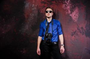 Studio portrait of stylish professional photographer man with camera, wear on blue shirt and necktie, sunglasses. photo