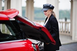 retrato de una hermosa modelo rubia de moda sexy con gorra y todo negro con maquillaje brillante cerca del auto rojo de la ciudad puso su bolso en el baúl. foto
