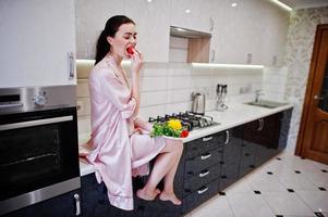 retrato de una bella joven vestida de rosa posando con un tomate en su cocina. foto