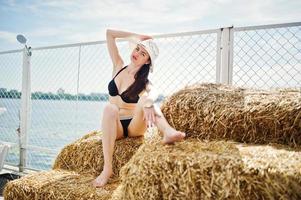 Portrait of a gorgeous girl in black bikini swimsuit posing on the hay bale with a hat by the lake. photo