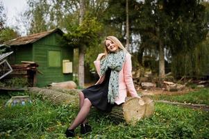 Young blonde girl at pink coat posed on autumn park, sitting on cut tree against house of the forester. photo