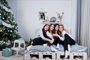 cuatro lindas amigas usan suéteres cálidos y pantalones negros en la cama en la habitación decorada de año nuevo en el estudio. foto