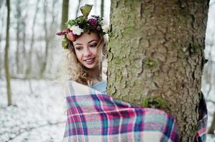 Curly cute blonde girl with wreath in checkered plaid at snowy forest in winter day. photo