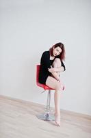 Red haired girl on black dress tunic sitting on red chair against white wall at empty room. photo