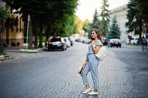 Portrait of a beautiful model in striped overall posing with hat and a backpack on a street with trees in a town. photo