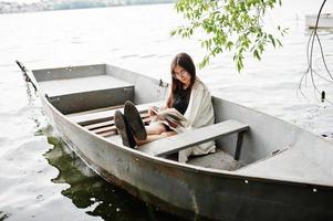 Portrait of an attractive woman wearing black polka dots dress, white shawl and glasses reading a book in a boat on a lake. photo