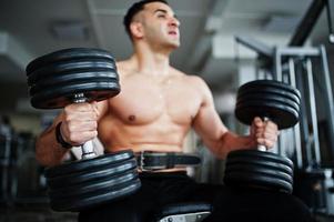 Muscular arab man training in with dumbbells modern gym. Fitness arabian men with naked torso doing workout . photo
