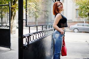 chica pelirroja con gafas con bolso rojo posado en la calle de la ciudad. foto