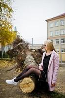 Blonde girl at glasses and pink coat, black tunic sitting on cut tree. photo