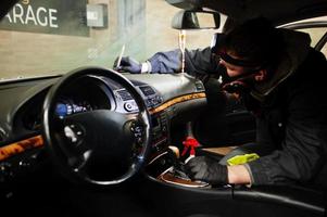 hombre en uniforme y respirador, trabajador del centro de lavado de autos, limpieza del cepillo de limpieza interior del auto. concepto de detalle de coche. foto