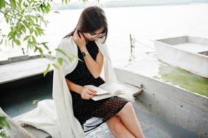 Portrait of an attractive woman wearing black polka dots dress, white shawl and glasses reading a book in a boat on a lake. photo