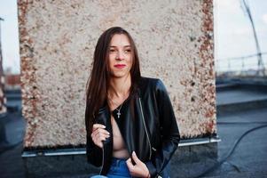 retrato de una hermosa joven con chaqueta de cuero negro, jeans y zapatillas sentadas en una azotea. foto