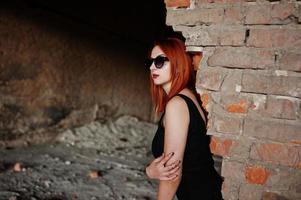 Red haired stylish girl in sunglasses wear in black, against abadoned place with brick walls. photo