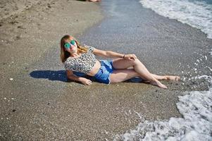 Beautiful model relaxing on a beach of sea, wearing on jeans short, leopard shirt and sunglasses. photo