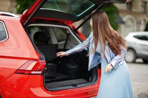 Outdoor photo of gorgeous woman posing near orange suv car with open car trunk.