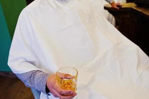 Customer holding a glass of whiskey sits in a chair in barbershop. photo