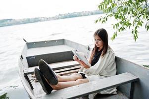 Portrait of an attractive woman wearing black polka dots dress, white shawl and glasses reading a book in a boat on a lake. photo