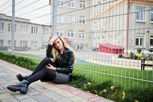 Blonde fashionable girl in long black leather coat against iron fence at street. photo