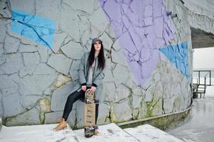 Stylish brunette girl in gray cap, casual street style with skate board on winter day against colored wall. photo