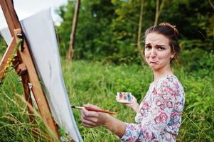 Portrait of a gorgeous sad young woman in beautiful dress sitting on the grass and painting on paper with watercolors. photo