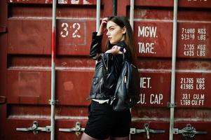 Stylish brunette girl wear on leather jacket and shorts with backpack against shipping container. photo