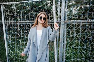 Girl in gray coat with sunglasses at small street stadium football gates. photo