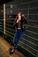 Night portrait of girl model wear on jeans and leather jacket against iron fence. photo