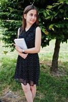 Portrait of a shy young woman in black polka dot dress holding books in the park. photo