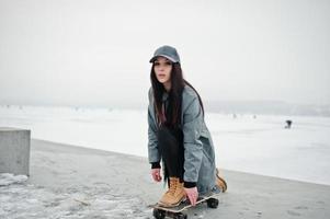 Stylish brunette girl in gray cap, casual street style with skate board on winter day. photo