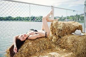 Portrait of a gorgeous girl in black bikini swimsuit posing on the hay bale with a hat by the lake. photo