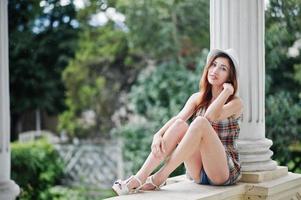 Increíbles piernas largas con tacones altos, ropa de niña con sombrero sentado cerca de columnas de edificios antiguos. foto