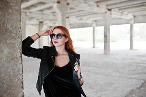 Red haired stylish girl in sunglasses wear in black, against abadoned place. photo